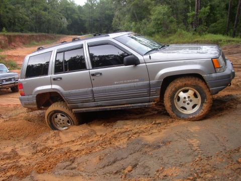 1991 Ford Bronco - A silver Jeep Grand Cherokee is stuck in thick mud on a dirt road, with one wheel deep in the mire.