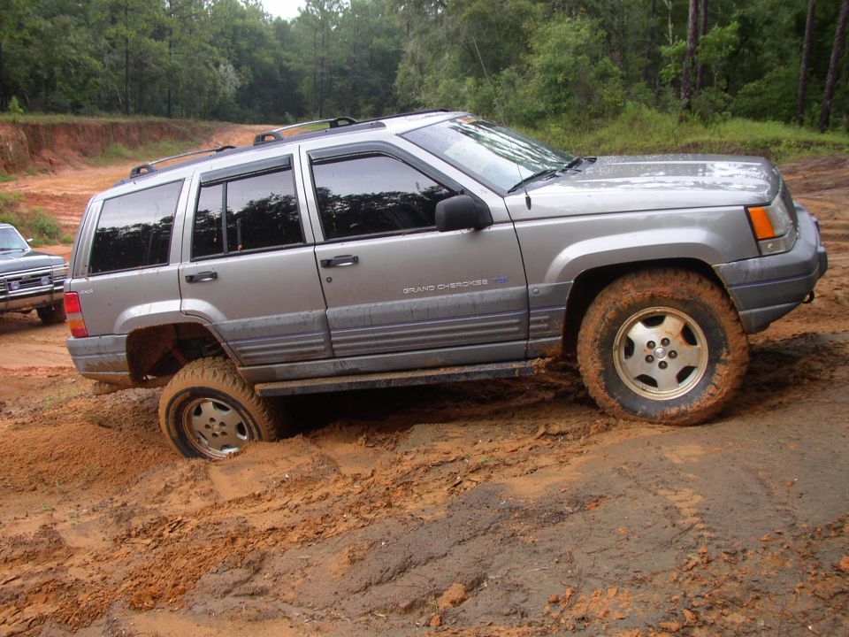 Silver Jeep Grand Cherokee stuck in mud, front wheel submerged. Another vehicle visible to the left