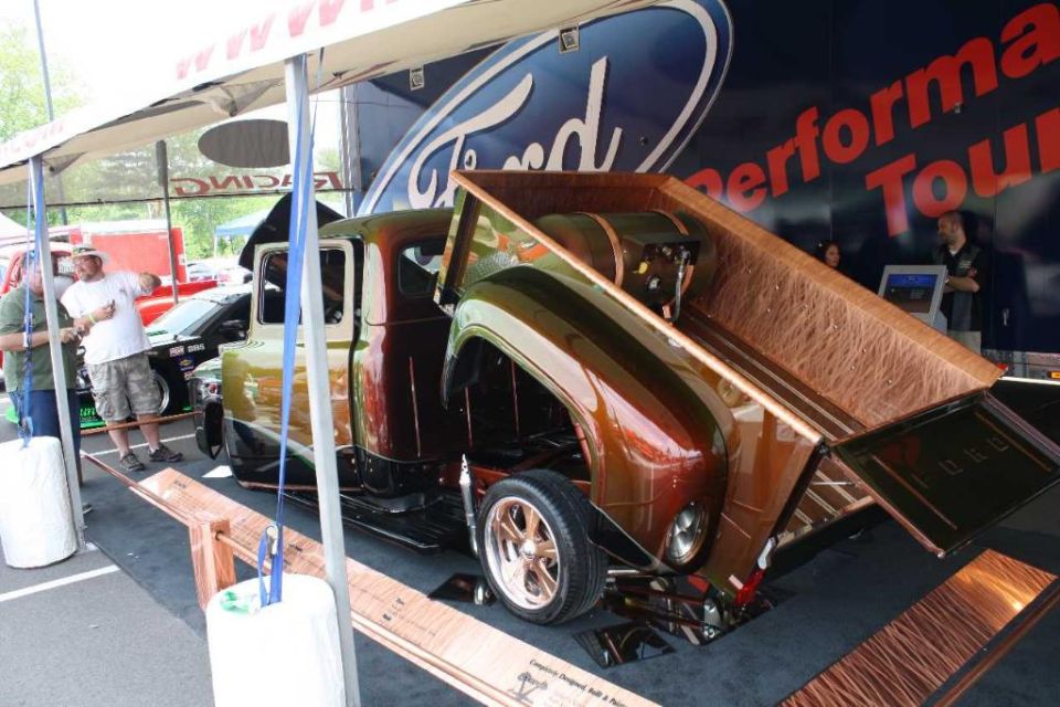 customized 1956 Ford F-100 pickup with a wood-lined bed and a metallic brown and green paint job, parked under a tent with a Ford Performance Tour banner