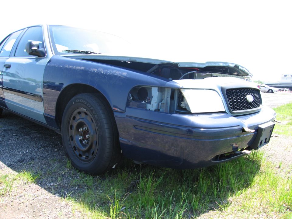 Blue Ford Crown Victoria with hood open, missing headlight, and steel wheels, parked on grass