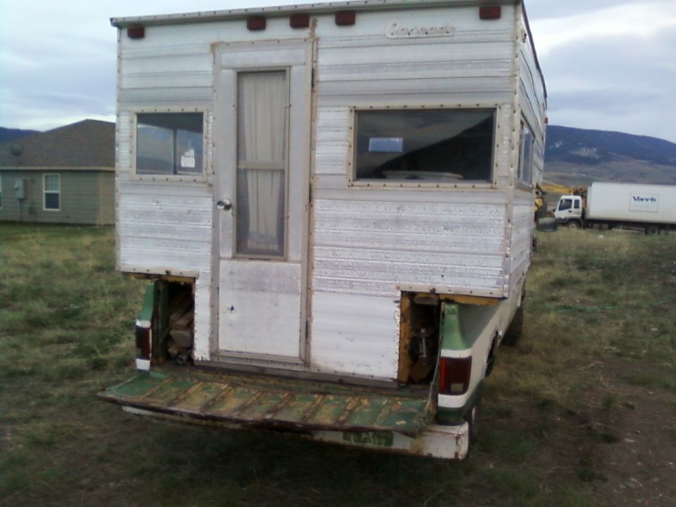 Camper on a green pickup truck bed, rear view. White camper body with windows and a door. Green tailgate is down