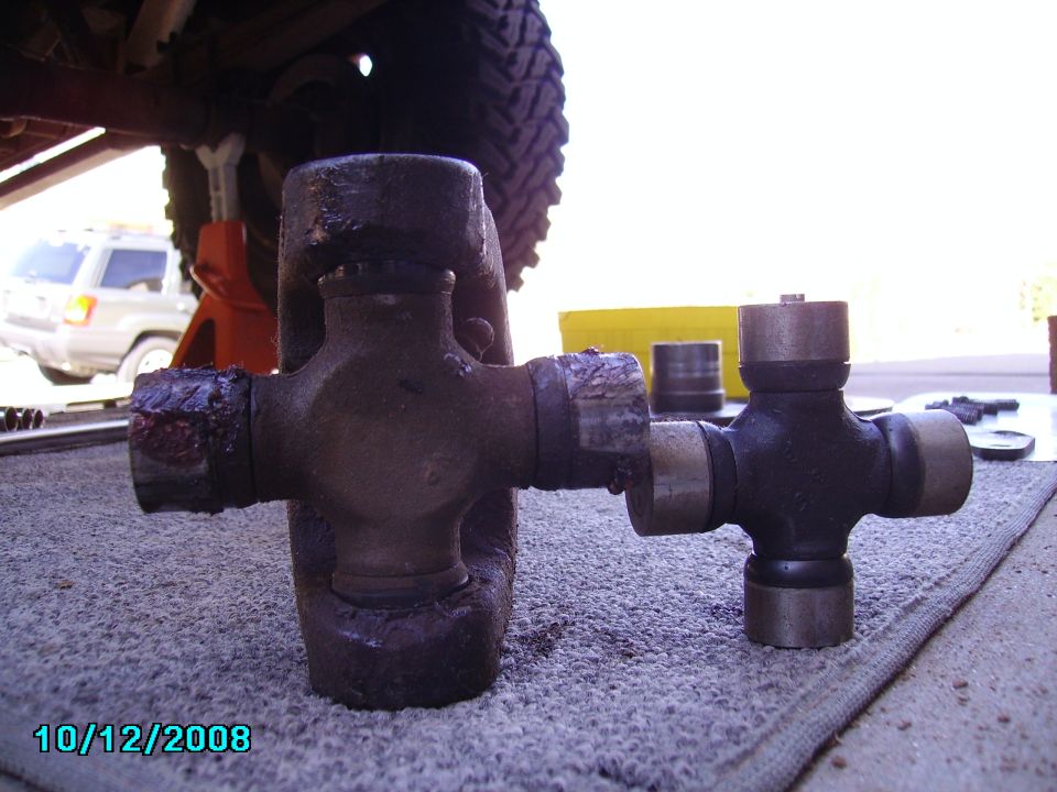 Two U-joint crosses compared side by side on carpet under a lifted 4x4 on jack stands