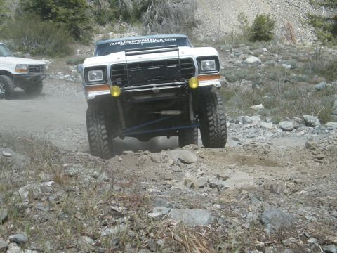 1982 Ford Bronco - Two Ford SUVs navigate a dusty, rocky mountain path, kicking up dust as they go.