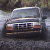 1993 Ford Bronco - A small, low-resolution signature-sized photo of a dark full-size Ford Bronco pushing through a deep muddy water hole, spraying brown water…