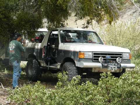 1982 Ford Bronco - A person photographs a lifted Ford Bronco parked in a wooded area with off-road modifications.