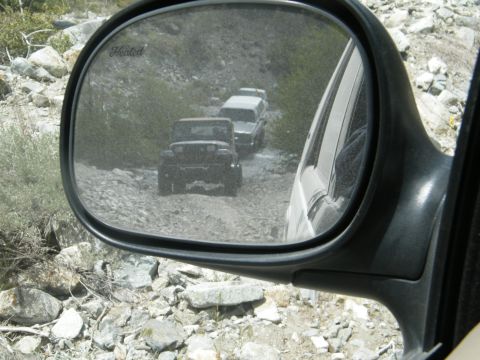 1982 Ford Bronco - Multiple Jeeps navigate a rough, rocky terrain on a dirt road, as seen through a side mirror.