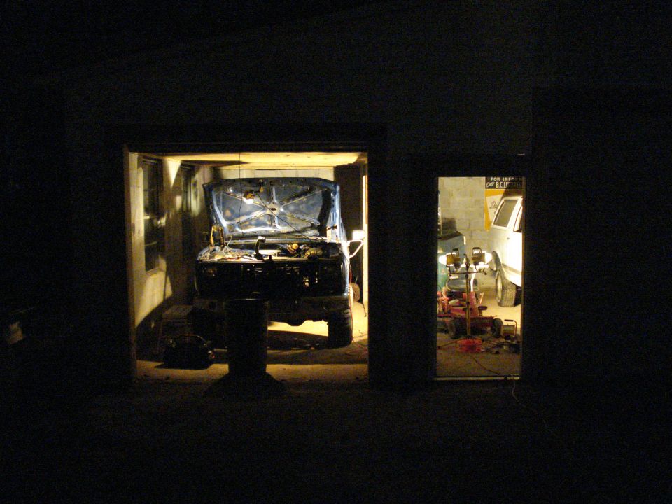 Blue truck with hood open, front view, in a garage. Another white vehicle is visible in an adjacent bay