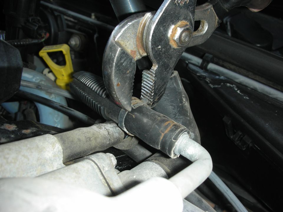 Pliers gripping a hose clamp on a black ribbed hose near engine components