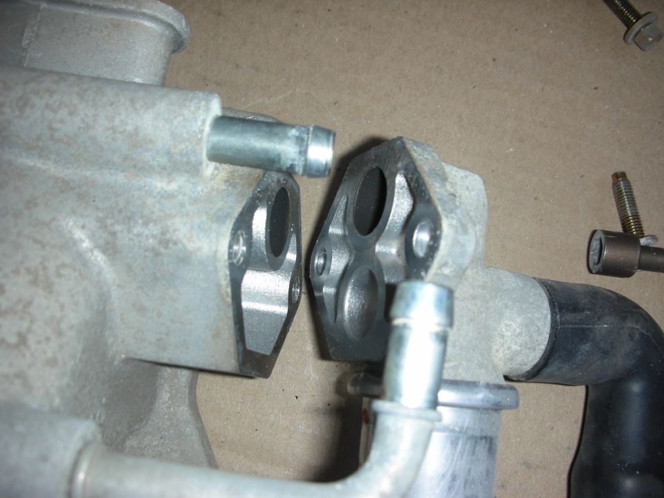Close-up of two metal engine parts, likely intake manifold components, with hoses attached. One part has a flange with four bolt holes