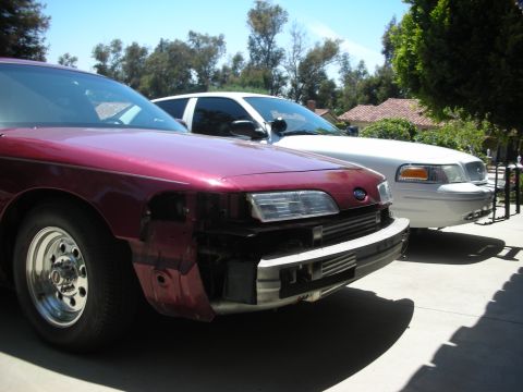 2004 Ford Crown Victoria - Two Ford Crown Victorias are parked outside on a sunny day, one with significant front-end damage.