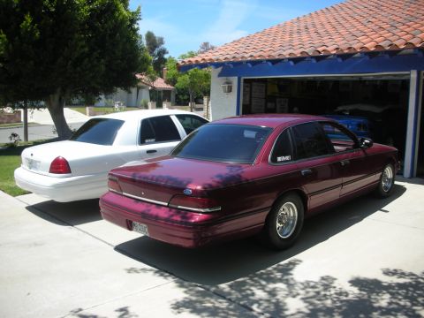 2004 Ford Crown Victoria - Two Ford Crown Victorias, one maroon and one white, are parked in a driveway on a sunny day.