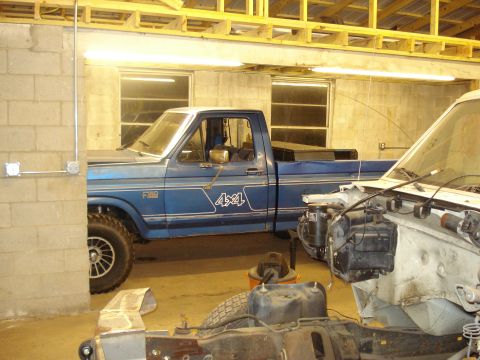 1967 Ford F-250 - A blue Ford F-150 pickup truck, likely from the 1980s, is parked inside a garage next to another vehicle undergoing repairs.
