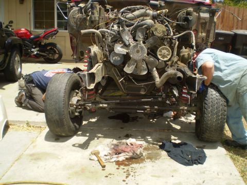 1969 Ford F-250 - Two mechanics work on a disassembled truck chassis with its engine exposed, while a red motorcycle is parked to the side.