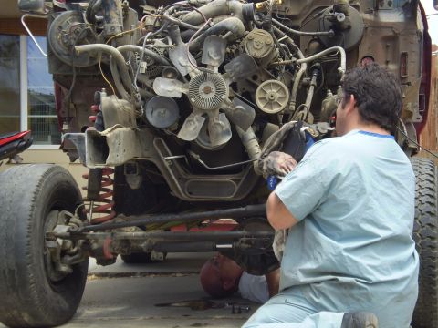 1969 Ford F-250 - Two mechanics are working on a heavily disassembled truck engine bay, with the front axle and suspension visible.