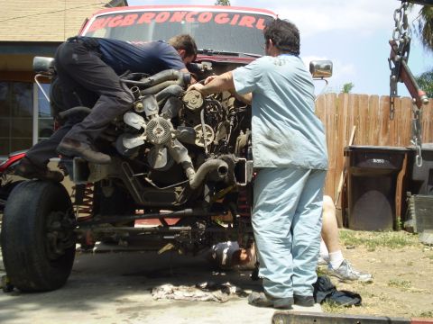 1969 Ford F-250 - Three people work on removing the engine from a red truck cab with a "BIGBRONCOGIRL" sticker.