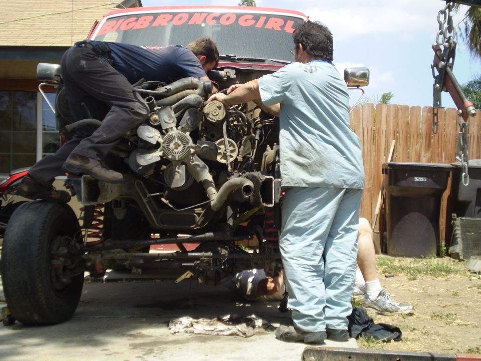 Red truck cab with "BIGBRONCOGIRL" sticker, engine being removed by three people