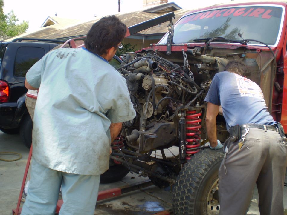 Two guys working on removing an engine from a red truck. A black SUV is parked behind it