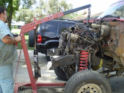 1969 Ford F-250 - A mechanic uses a cherry picker to remove the engine from a truck, with a black SUV parked behind.