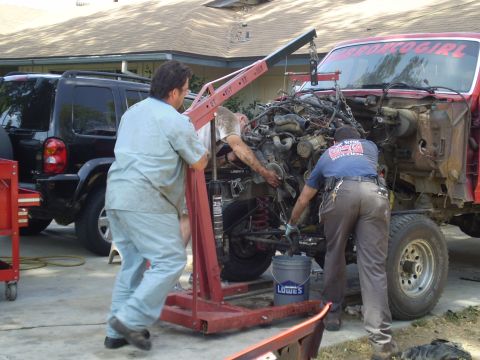 1969 Ford F-250 - Mechanics use an engine hoist to remove the engine from a red truck. A black SUV is parked nearby.