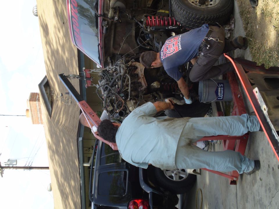 Two guys working on removing an engine from a red truck with an engine hoist. Another SUV is parked behind it