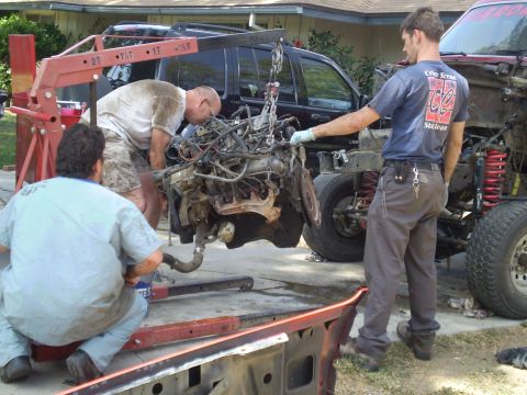 1969 Ford F-250 - Mechanics work on removing an engine from a truck using an engine hoist. Another vehicle is visible in the background.