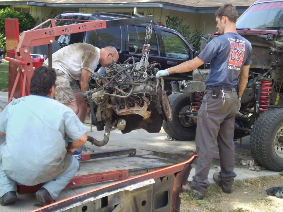 Engine hoist lifting an engine out of a red truck chassis; another truck is parked behind