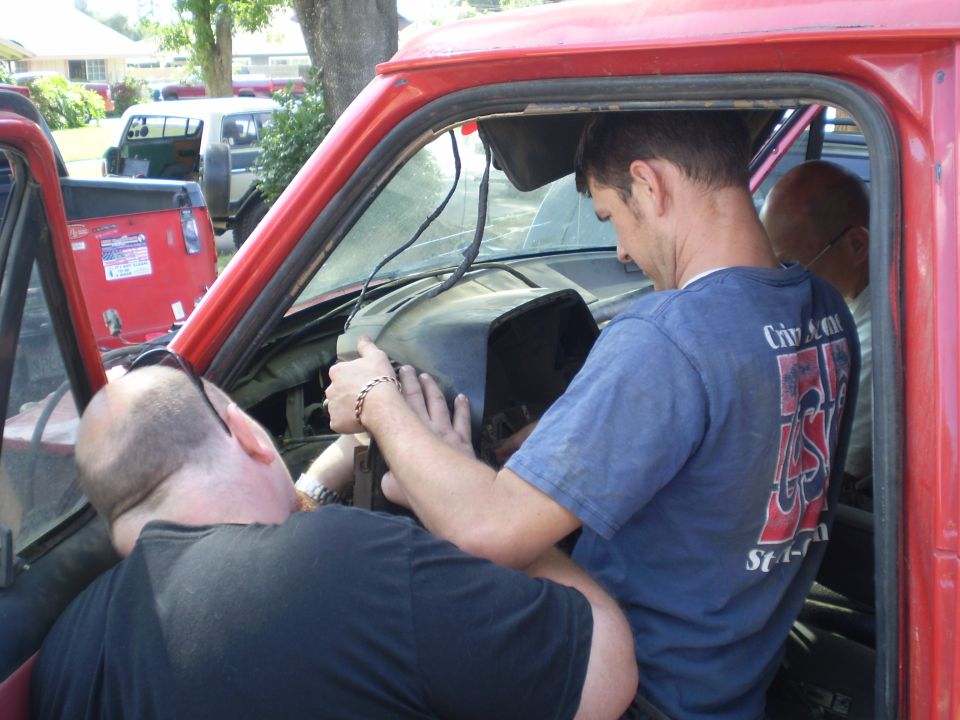 Two men working on the dashboard of a red truck. Another truck is visible in the background