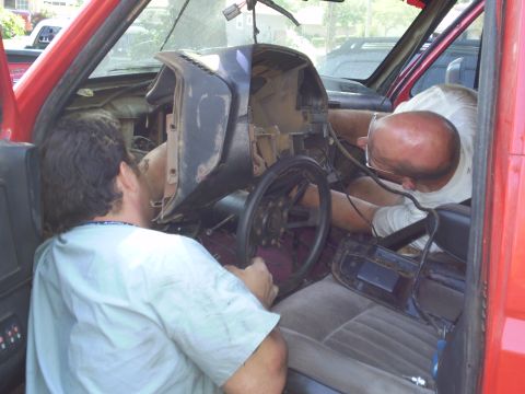 1969 Ford F-250 - Two people leaning into a red pickup cab repairing the dash and steering column with parts and tools scattered inside.