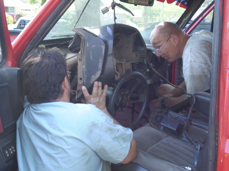Two men are working on the dashboard of a red vehicle. The dashboard is partially disassembled