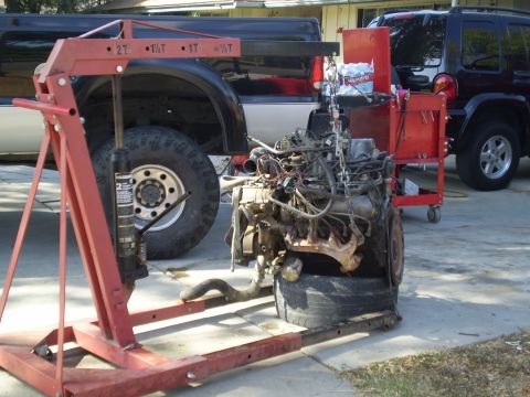 1969 Ford F-250 - An engine is suspended by a hoist, with parts of two vehicles visible behind it.