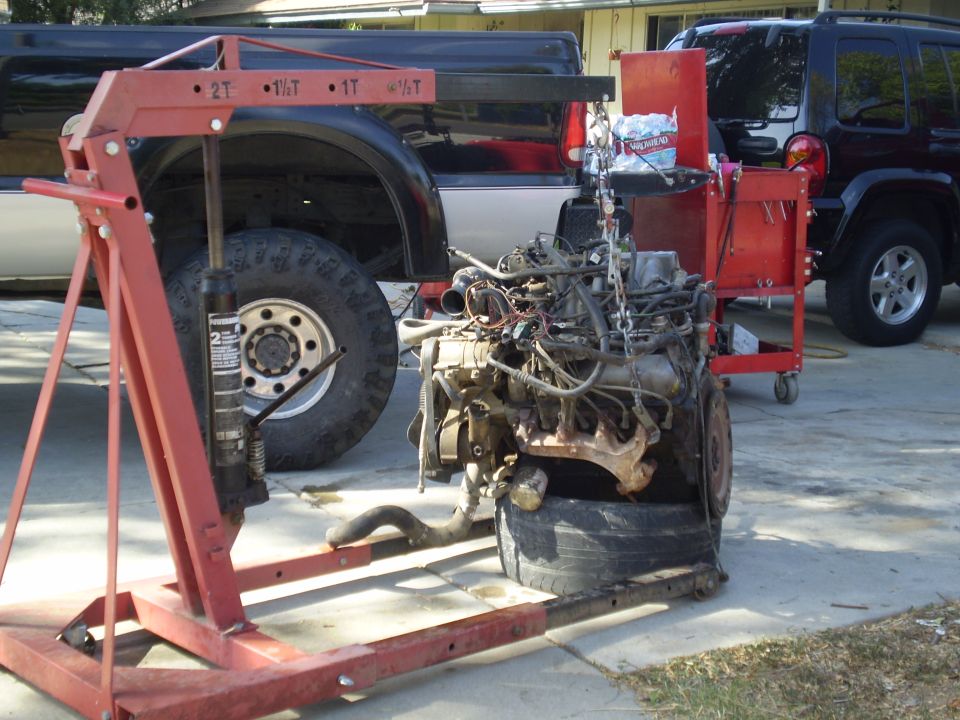 Engine hoist over a removed engine, with a pickup truck and SUV in the background