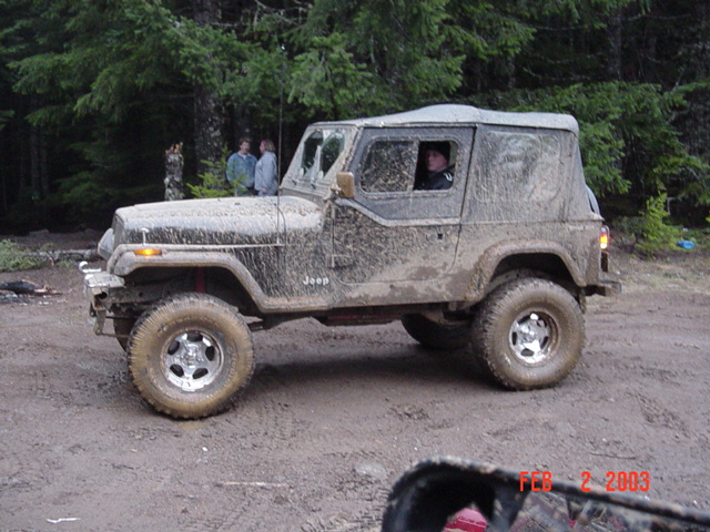 Side view of a mud-covered Jeep Wrangler YJ on a dirt lot with people standing in the background