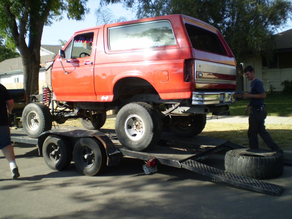 Red Ford Bronco on a trailer, lifted with large tires. A man is standing near the rear