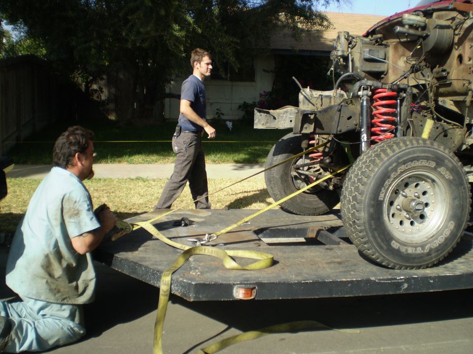 Truck chassis with red springs and front tires on a flatbed trailer, being secured by two men