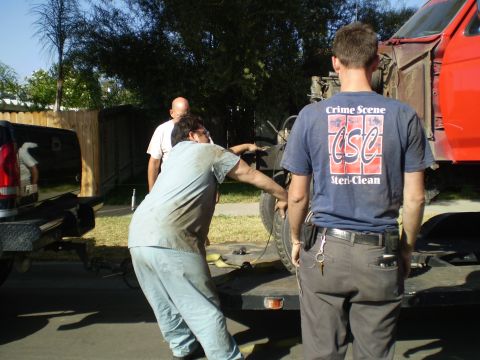 1969 Ford F-250 - Three men work to load a red truck onto a trailer, with one man wearing a shirt that says "Crime Scene Steri-Clean."