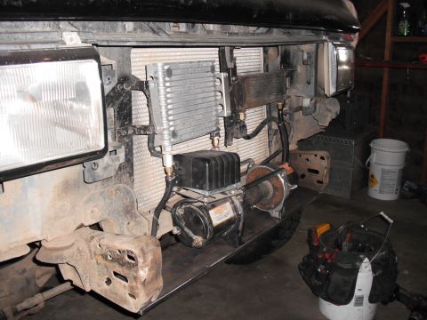 1991 Ford Bronco - Close-up of a winch and radiator assembly, with visible hoses and a black box, mounted on a metal plate.