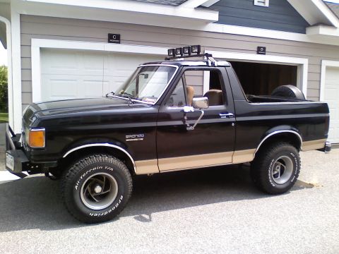 1990 Ford Bronco - Side view of a black and tan 1990 Ford Bronco parked in front of a garage, equipped with roof-mounted lights and an external spare tire.