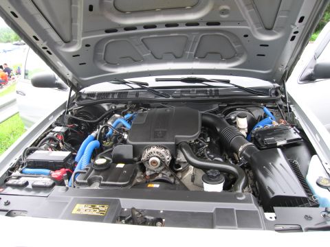 1991 Ford LTD Crown Victoria - Close-up of an open engine bay showing the V8 motor with blue spark plug wires, accessory belt and air intake under a raised silver hood.