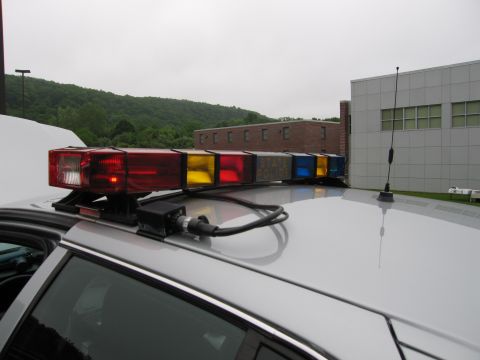 1991 Ford LTD Crown Victoria - Close-up of a police light bar with red, amber and blue lenses and a whip antenna mounted on a white cruiser roof, with buildings behind.