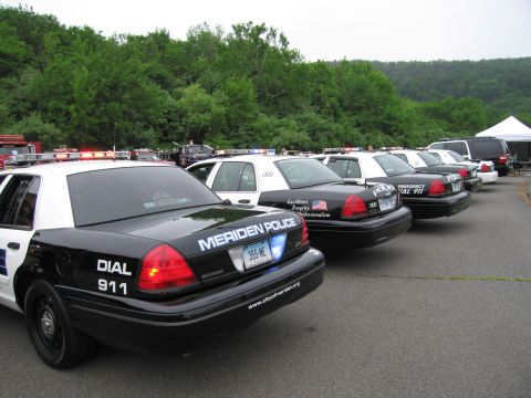 1991 Ford LTD Crown Victoria - angled row of black-and-white Meriden Police cruisers and other emergency vehicles parked in a lot with a tent and woods behind.