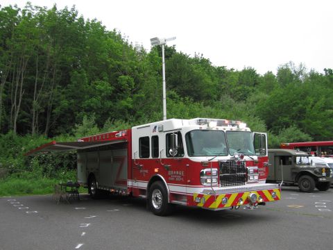 1991 Ford LTD Crown Victoria - red and white Orange Fire Dept. rescue truck with a side compartment awning open, parked beside a military vehicle near woods.