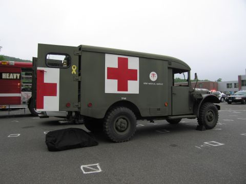 1991 Ford LTD Crown Victoria - Side view of an olive-drab U.S. Army medical service ambulance truck displaying a large red cross, with a cover bundle on the ground beside…