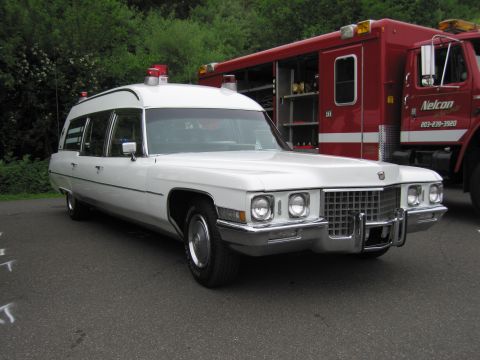1991 Ford LTD Crown Victoria - white Cadillac professional car styled as an ambulance/hearse with roof beacons, parked in front of a red fire apparatus.