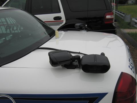 1991 Ford LTD Crown Victoria - Two black radar or camera units mounted on the white hood of a police cruiser, with a dark SUV behind.