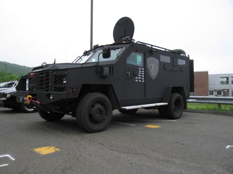 1991 Ford LTD Crown Victoria - black armored tactical police truck with roof turret and side gun ports, parked outdoors near a building.