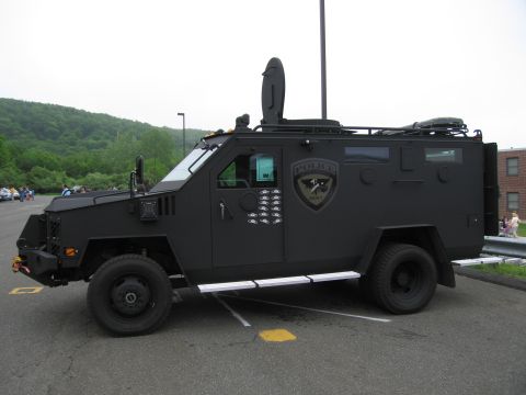 1991 Ford LTD Crown Victoria - Side profile of a black armored tactical police truck displaying a police crest, parked in a lot with hills behind.