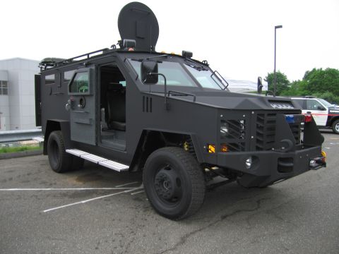 1991 Ford LTD Crown Victoria - black armored tactical police truck with its side door open and roof turret raised, parked near a building.