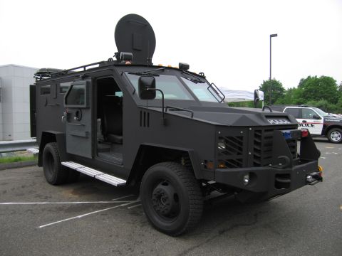1991 Ford LTD Crown Victoria - black armored tactical police truck with grille gun ports and open side door, parked beside a white-and-black cruiser.