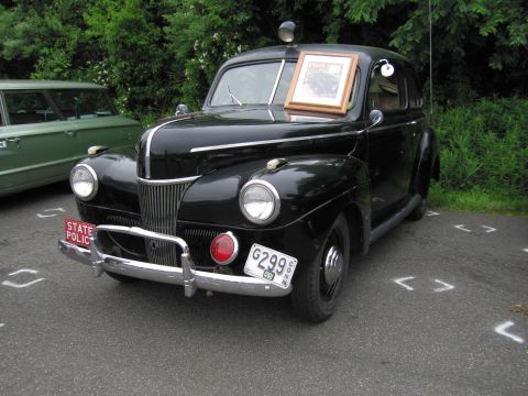 1991 Ford LTD Crown Victoria - black early-1940s Ford two-door sedan marked 'STATE POLICE' with a roof beacon, parked beside a green wagon.