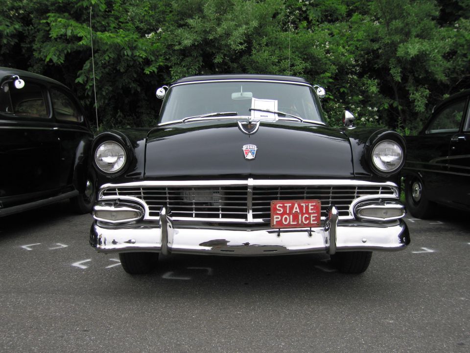 Front-on view of black 1950s Ford with chrome grille and red State Police plate